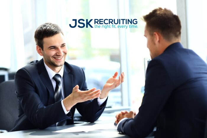 A job interviewer gestures and smiles at a job applicant across a desk.