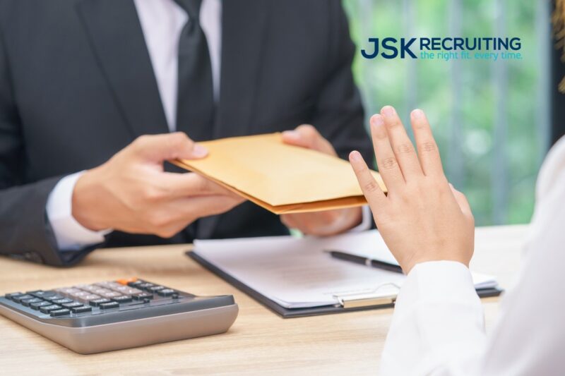 A businessman hands a sealed counteroffer to a job candidate, who holds up her hand to politely decline it. Photo is watermarked with the JSK Recruiting logo.