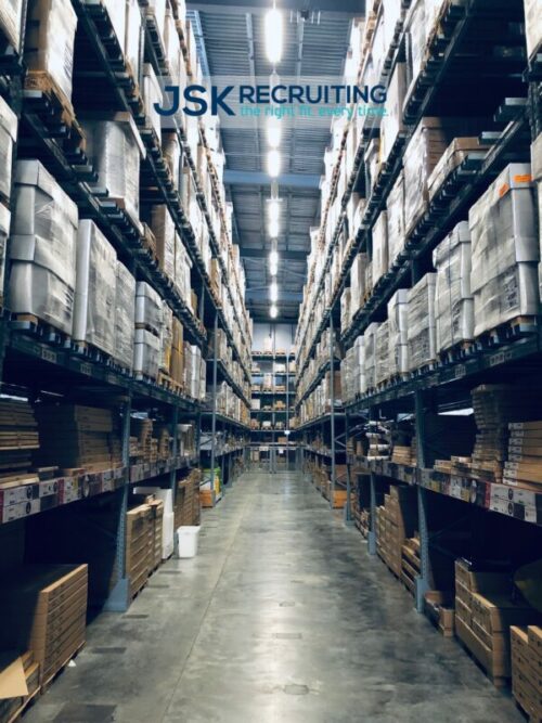 A view down an aisle in warehouse stacks loaded with inventory crates. Photo is watermarked with the JSK Recruiting logo.