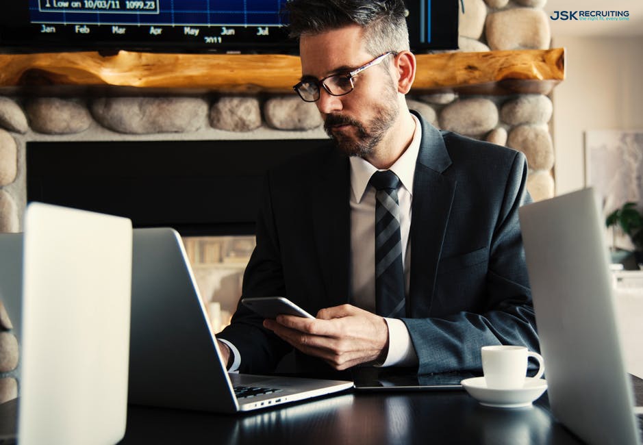 A young businessman in glasses reviews figures on his phone and laptop with a cup of coffee under a screen displaying job market figures.