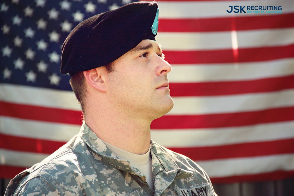 A US Army solider in fatigues and beret gazes upward in front of an American flag.