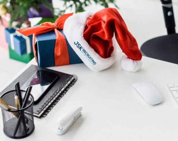 A Santa hat watermarked with the JSK Recruiting logo lays on top of a present at a business desk.