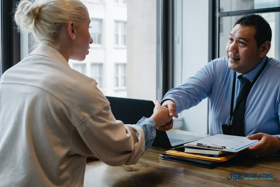 Two people shaking hands at a table in front of windows.