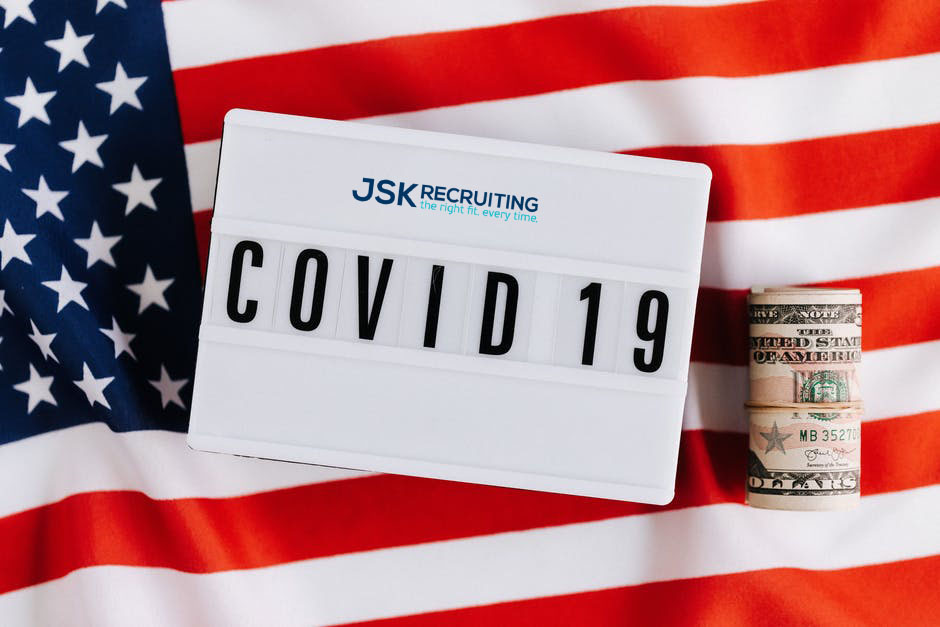 A JSK Recruiting sign that reads "COVID 19" sits on an American flag next to a roll of 5-dollar bills.