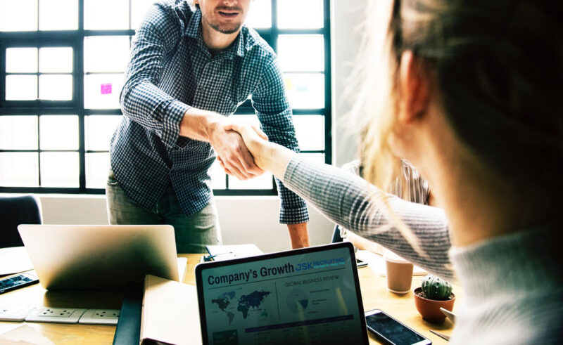 Two people shaking hands over a table with a laptop.