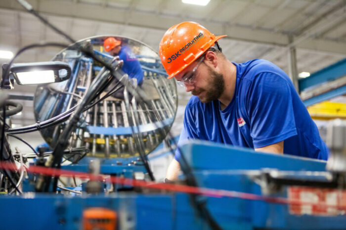 A worker in an orange hardhat watermarked with the JSK Recruiting logo operates a piece of heavy machinery next to a mirror in a manufacturing plant.