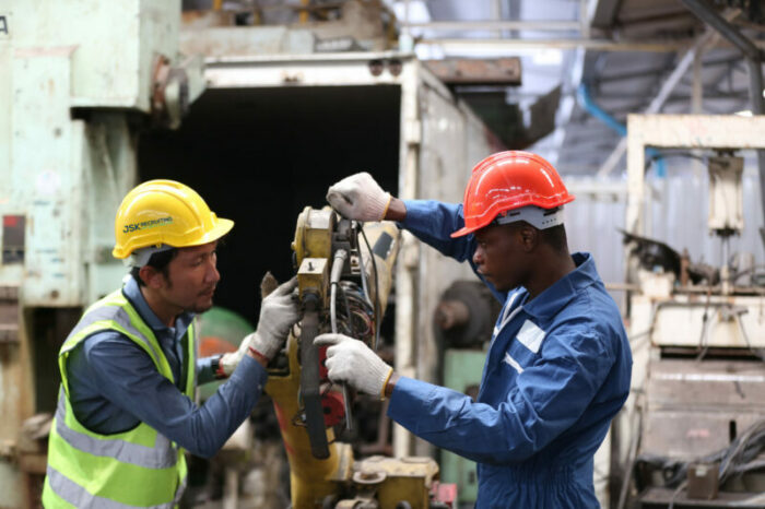 Two factory workers in safety vests and hardhats watermarked with the JSK Recruiting logo cooperate at a piece of heavy machinery.