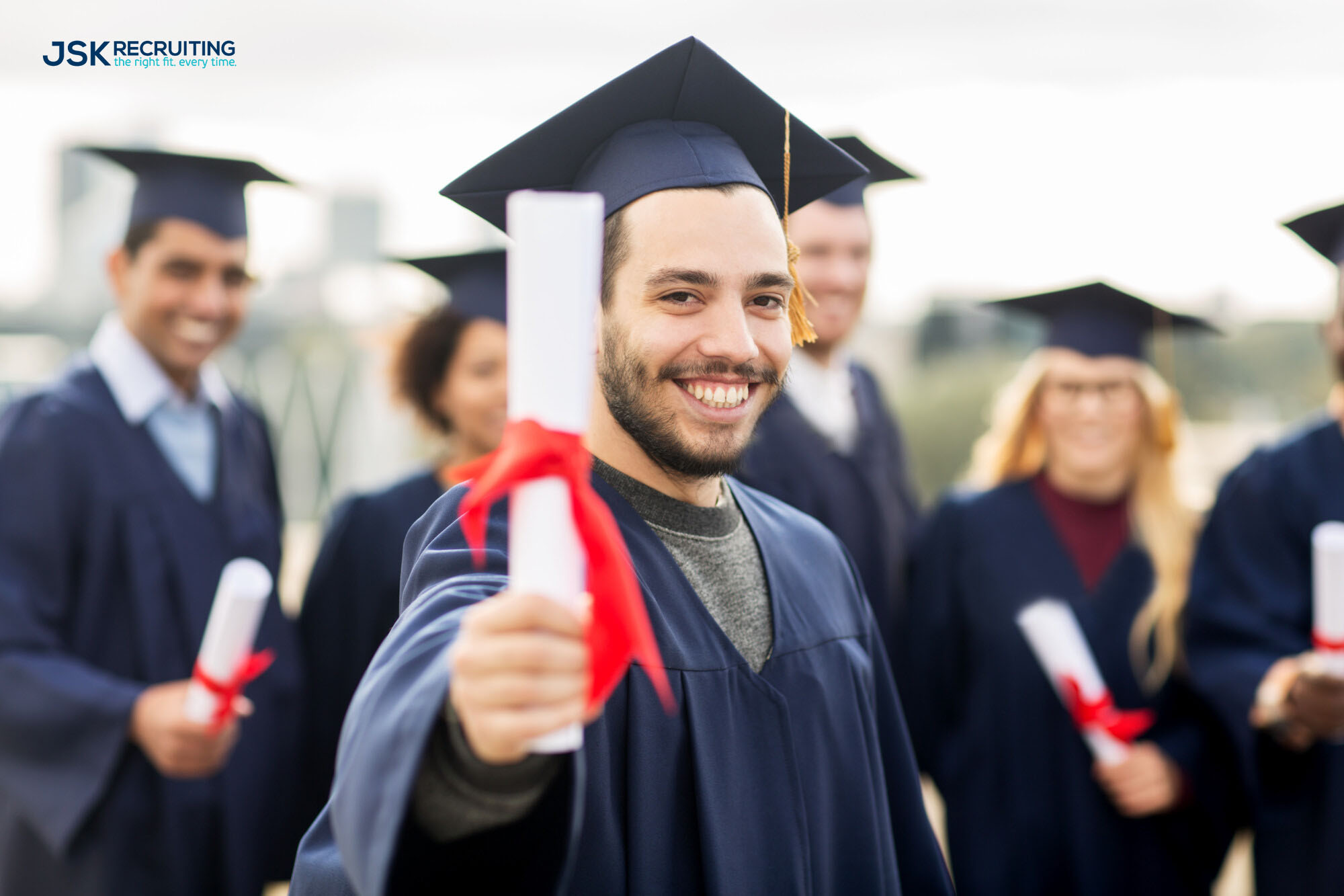 A young man in a cap and gown smiles as he presents his college degree. Photo watermarked with the JSK Recruiting logo.