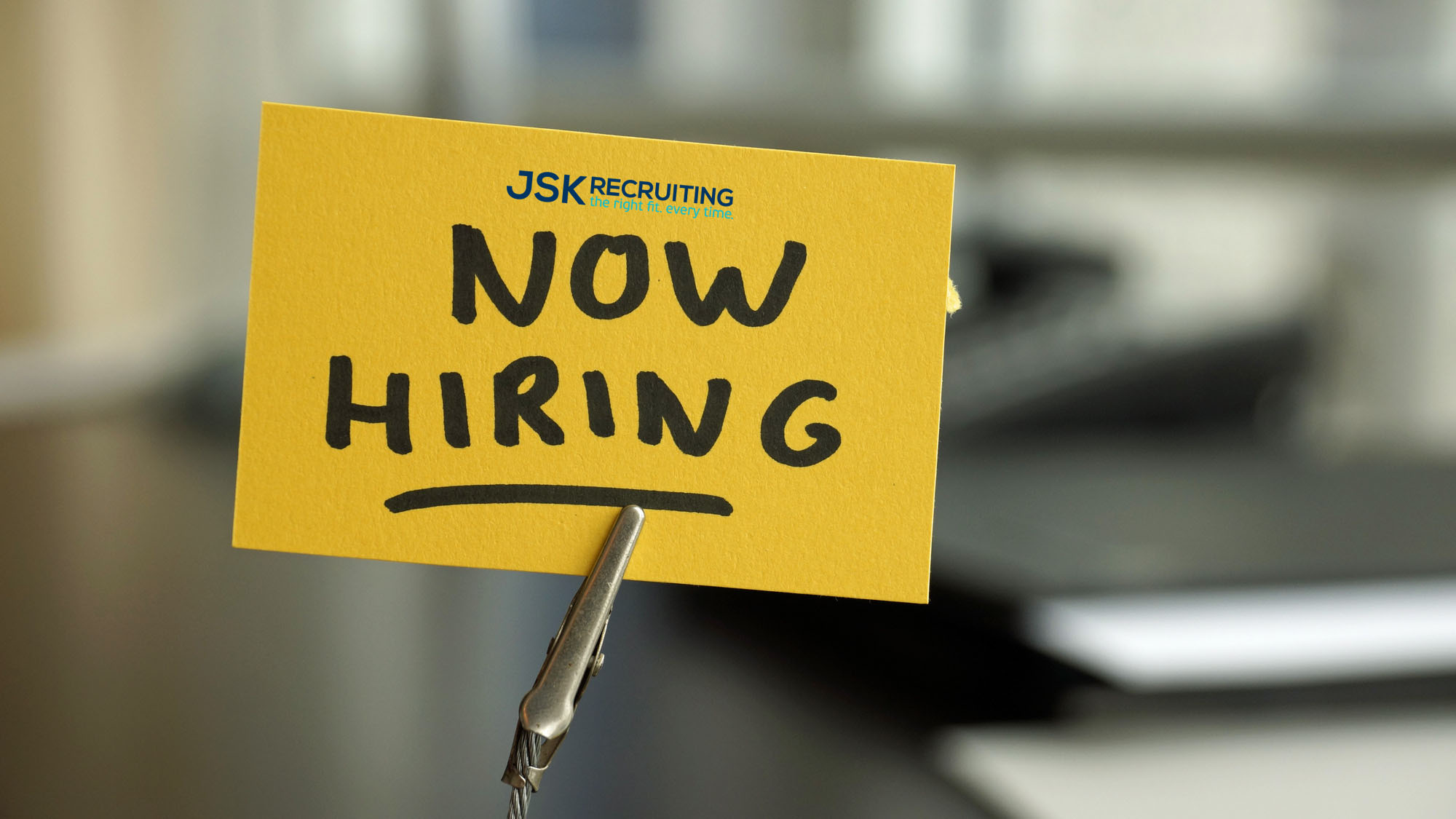 A yellow sign that reads "now hiring" on a clip emblazoned with the JSK Recruiting letterhead.