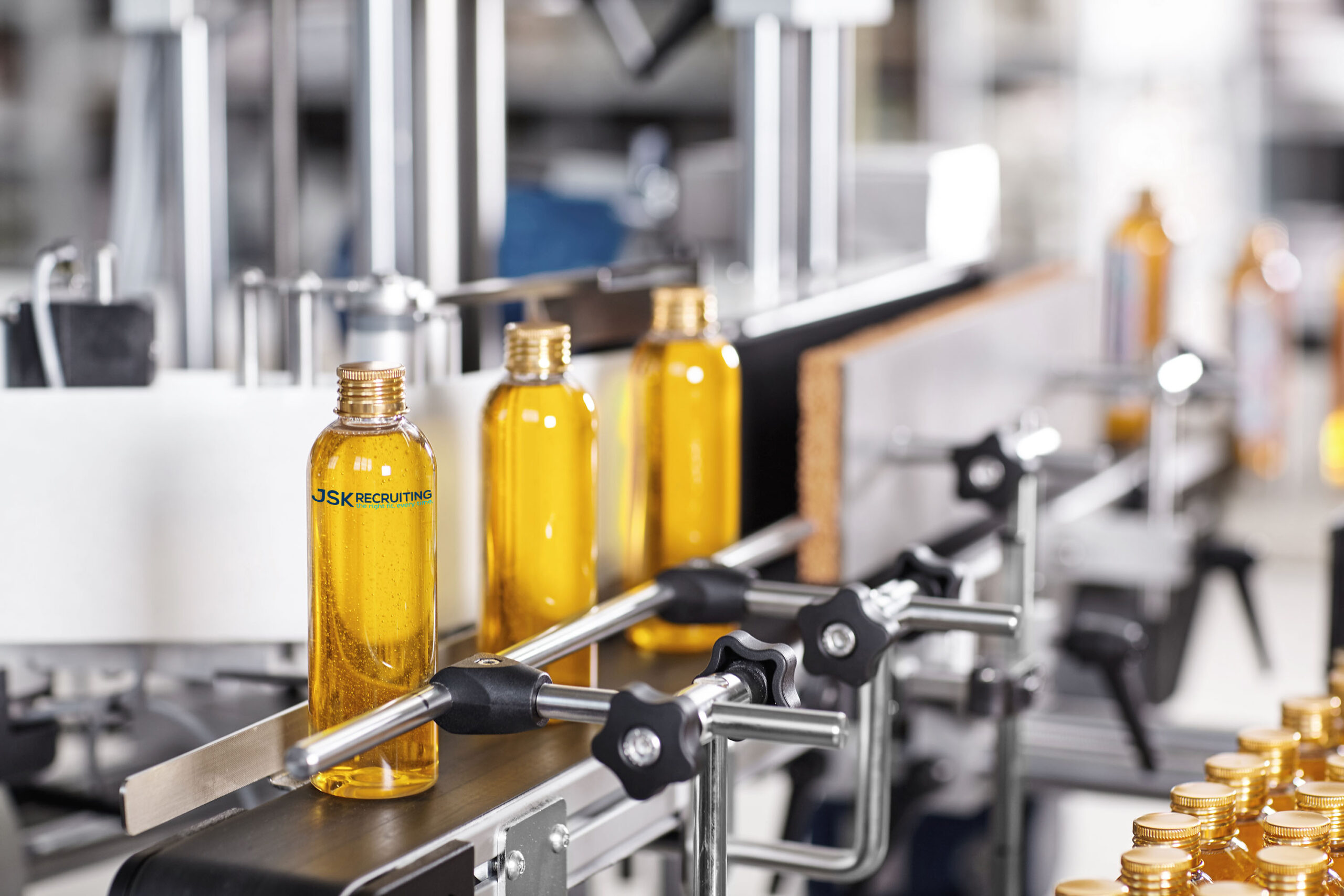 A line of yellow bottles emblazoned with the JSK Recruiting logo on a conveyor belt.