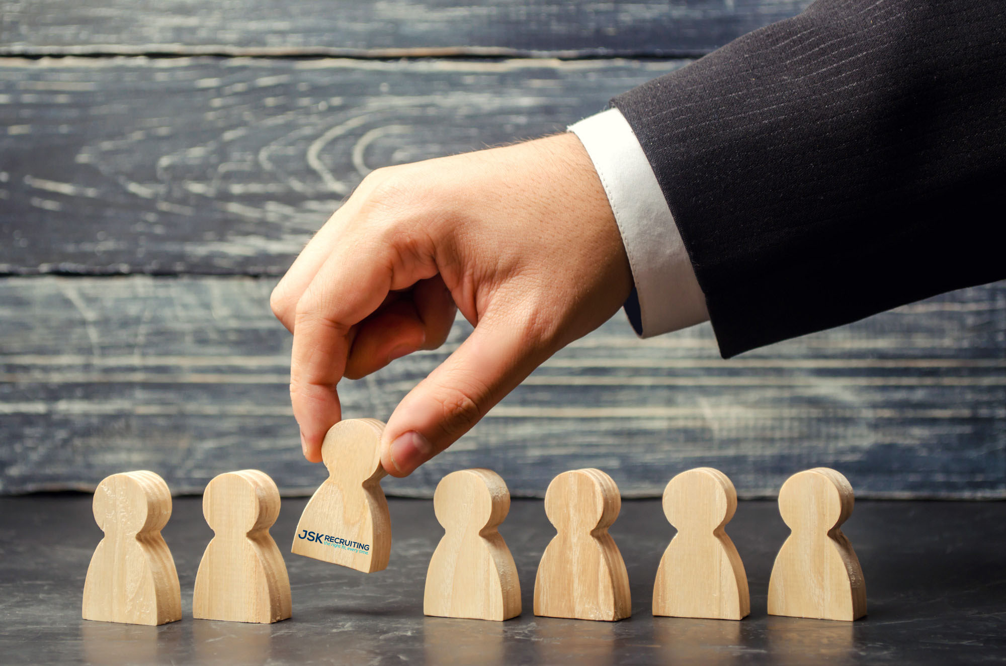 A man's hand chooses one wooden figure emblazoned with the JSK Recruiting logo from a row of similar blank wooden figures. A visual metaphor for being recruited for a job.