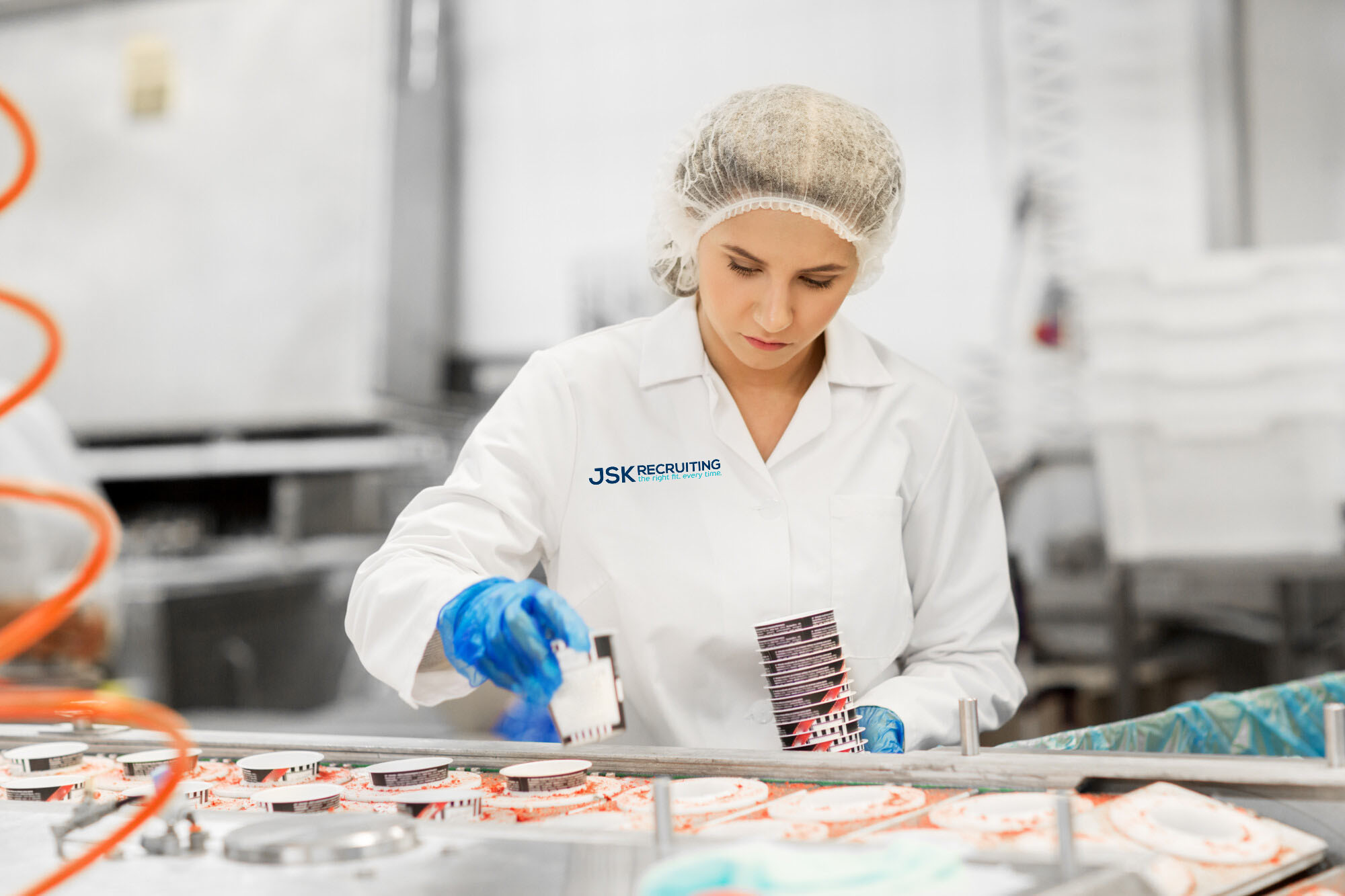 A woman in white lab coat emblazoned with the JSK Recruiting logo and blue gloves inspects ice cream containers on an assembly line.