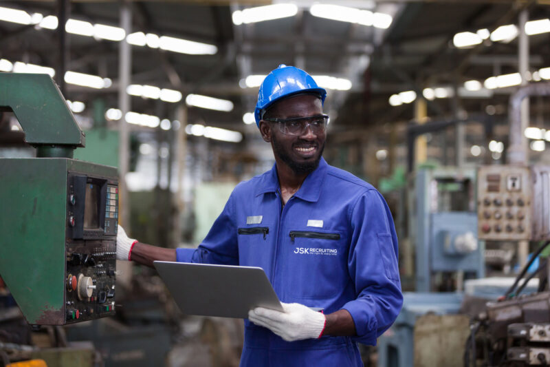 An African American factory mechanic in a blue hard hat, coveralls labeled with the JSK Recruiting logo and safety glasses smiles as he checks on a machine.