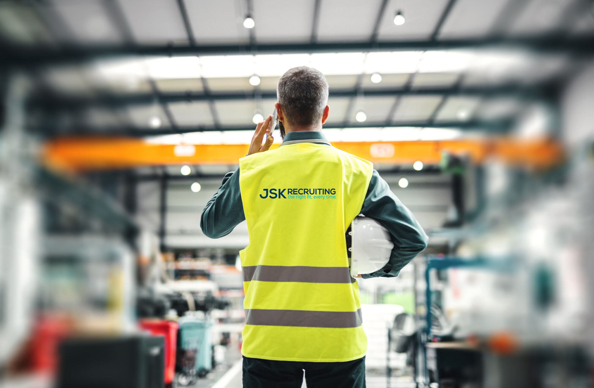 An industrial manager in a JSK Recruiting safety vest viewed from behind talks on a cellphone.