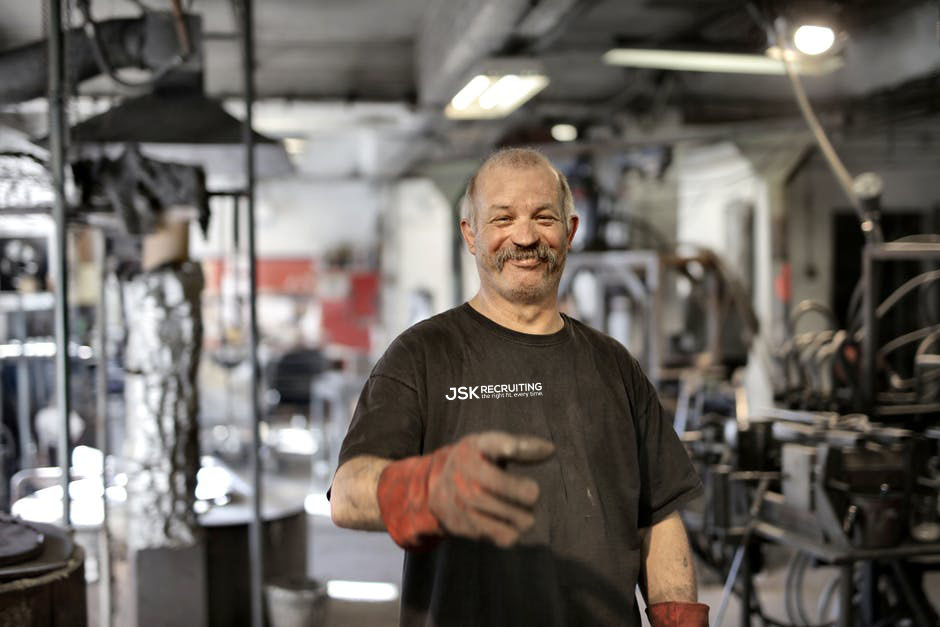 A mustachioed mechanic in gloves in a workshop smiles at the camera. His shirt is emblazoned with the JSK Recruiting logo.