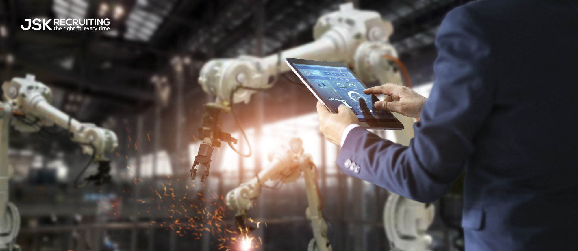 A man in a suit reviews data on a tablet in front of robotic arms welding with sparks in an intelligent factory.