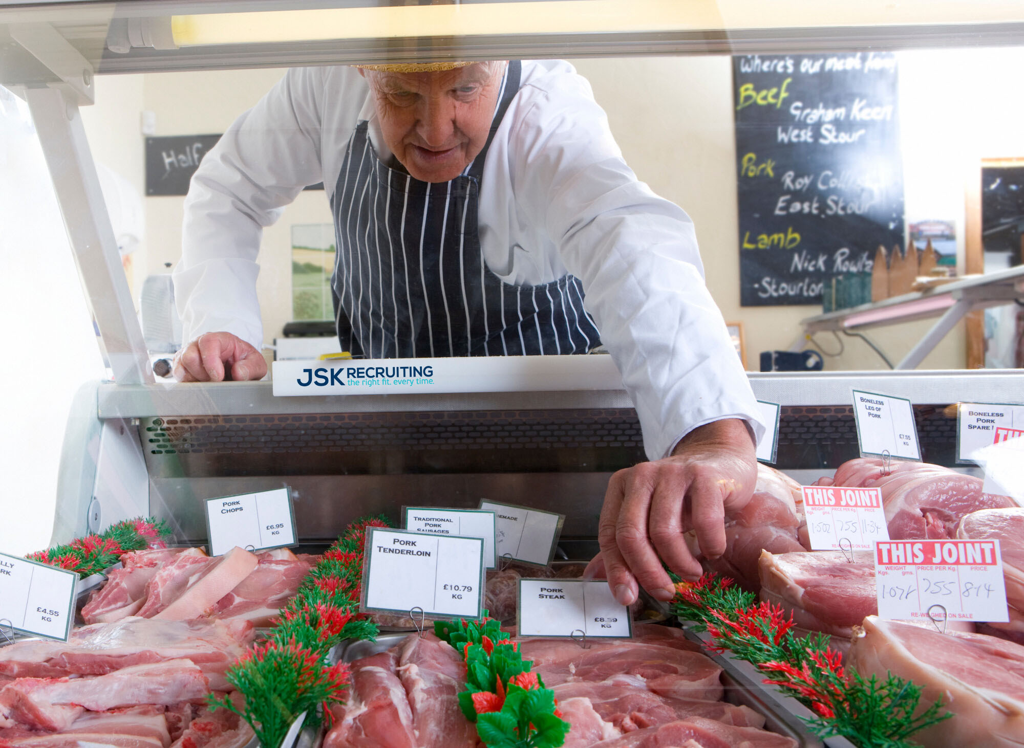 Older butcher in apron and hat arranging display of meat in shop display refrigerator emblazoned with the JSK Recruiting logo.