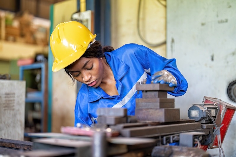 A young African American factory worker in a hard hat and coveralls concentrates as she operates machinery.