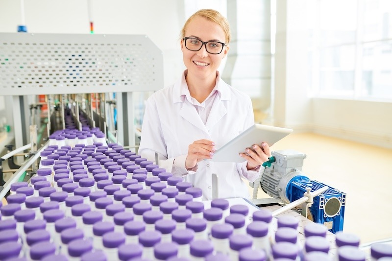 A smiling young female technologist in white lab coat holds tablet near purple containers on a conveyor.