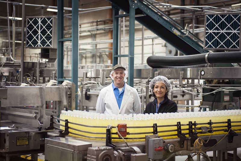 Two factory workers in protective gear smile next to a conveyor belt with rows of bottles with lemon-flavored water.